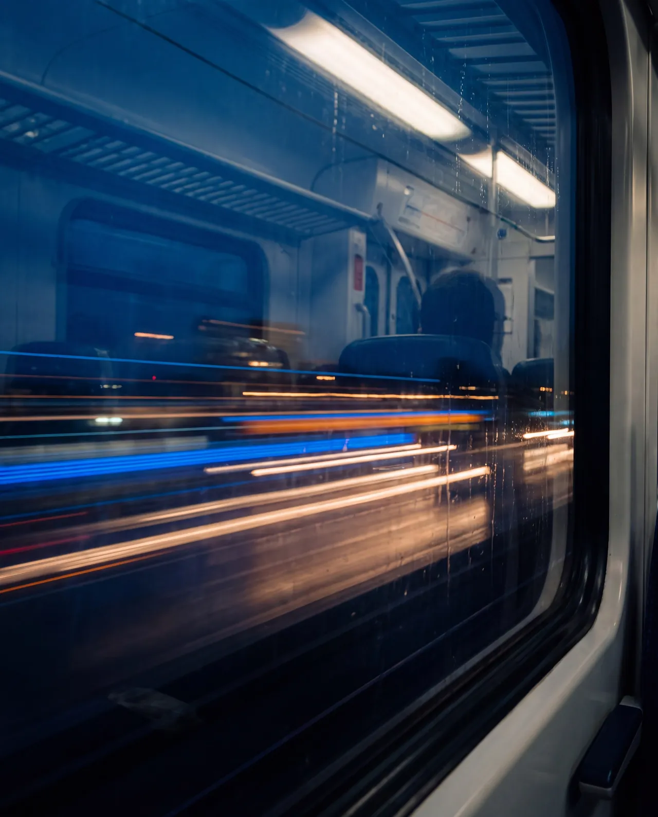 City lights streaking past a train window at night, with the carriage reflected in the glass.
