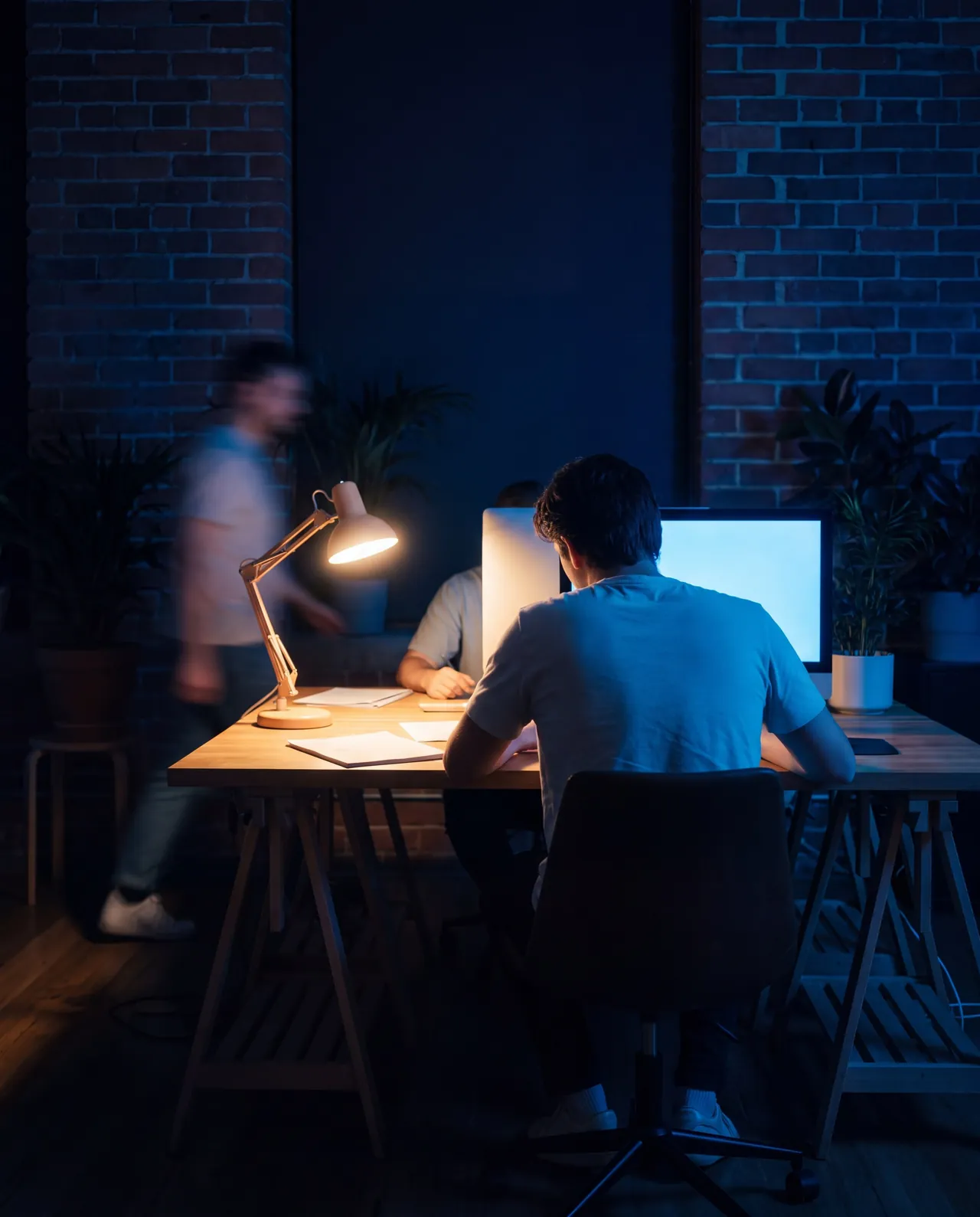Two people working late at a shared desk in a brick-walled studio, by a desk lamp and a monitor, as someone walks past.